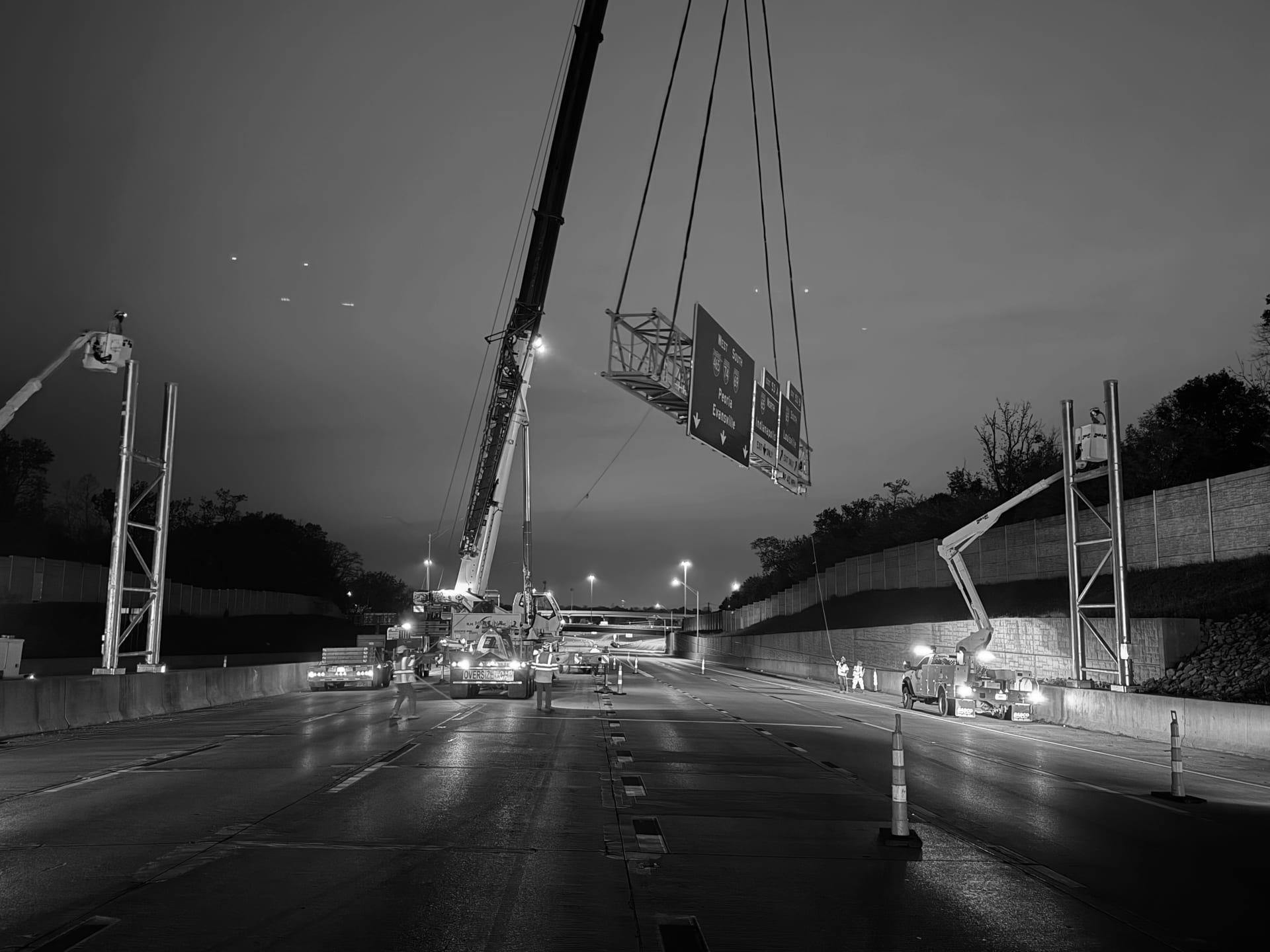 Nighttime erection of an overhead highway sign bridge with a crane lifting a multi-panel sign assembly above an I-465 interchange — James H Drew Corporation sign structure installation